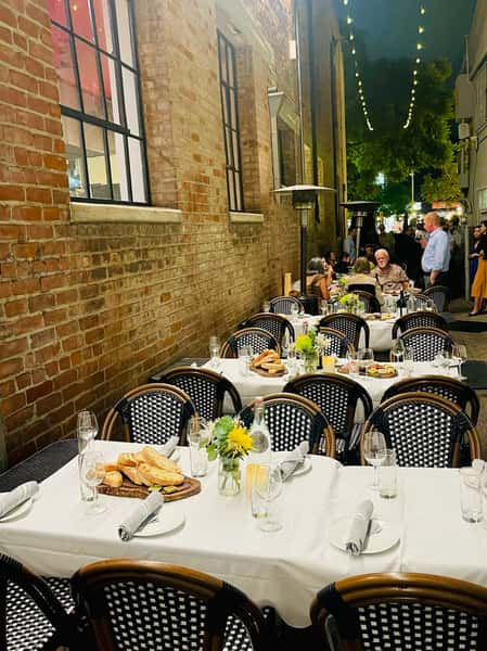 Charming outdoor dining area with wooden tables, elegant table settings, and vibrant flowers. Warm ambiance created by patio heaters and string lights. Perfect for gatherings.