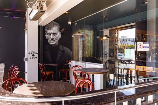Stylish bar interior featuring wooden tables, red chairs, and a striking mural of a man. The space is inviting, perfect for socializing and enjoying drinks in a modern setting.