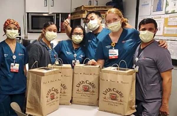 Group of six nurses and doctors wearing masks and blue scrubs standing behind 4 large Urth food bags on table. Male nurse in background is giving thumbs up.