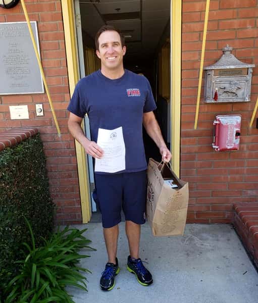 Santa Monica Fireman in blue T-shirt and shorts holding large Urth Caffé bag of lunch boxes. He is standing just outside a narrow doorway leading into the firestation.
