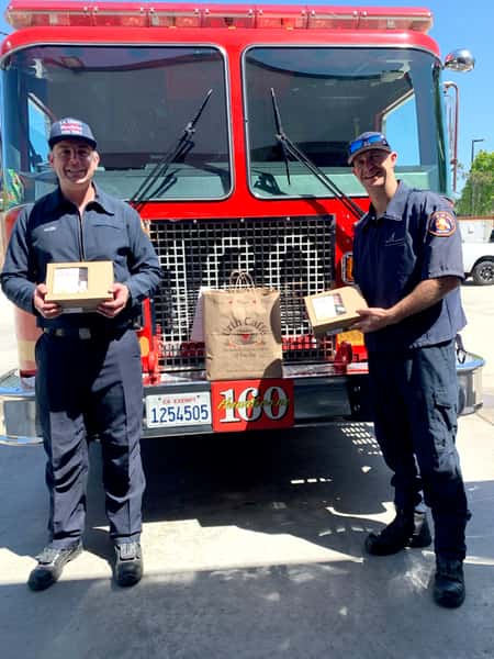 2 firemen holding Urth lunch boxes standing in front of red firetruck fr
