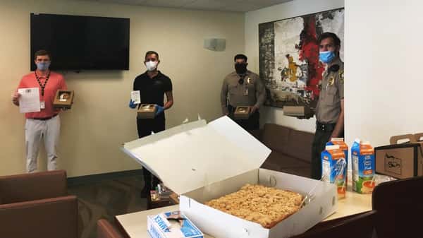 Room inside Beverly Hills City Hall with two officers and two staff member standing against walls. Table in foreground has large Urth Caffé crumb coffee cake, OJ cartons and paper coffee despensers.