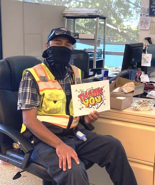 Man sitting at desk with black mask, hat, yellow vest and holding a thank you sign.