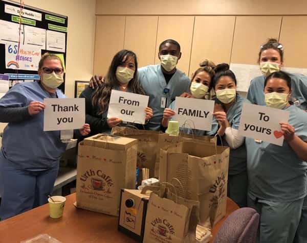 7 White Memorial Hospital nurses with yellow masks holding four sheets of paper that say "Thank You From our Family To Yours" with image of red heart. They are standing behind a table full of Urth Caffé food bags and coffee.