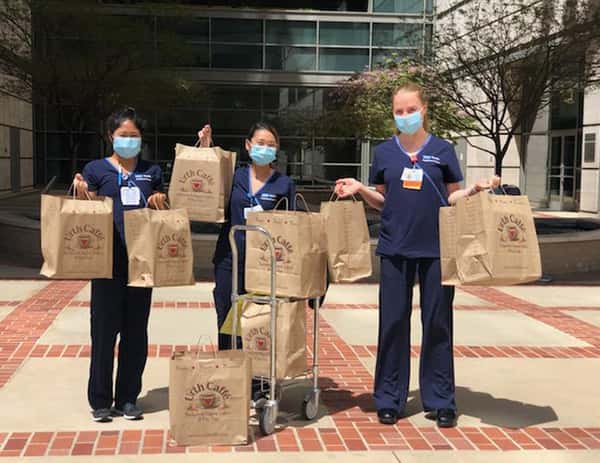 Three nurses with blue masks standing in front of the Ronald Reagan UCLA Med center holding multiple large Urth food bags and a cart with bags in hospital courtyard.