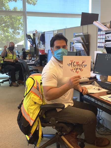 Man sitting at desk with mask and holding a thank you sign. In the background is another man is sitting at another desk with mask and holding the Urth Caffe "To Our Brave Heroes" card.