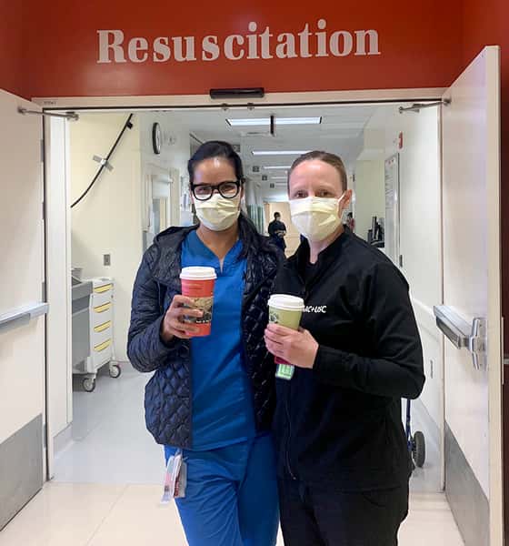 Two nurses with medical masks holding Urth Caffé To Go coffee cups. They are standing in front of open double doors with "Resuscitation" above them.
