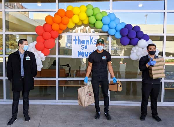 Three people standing in front of large glass window of St. Joseph's hospital with band of balloons and Urth Lunch boxes and bags.