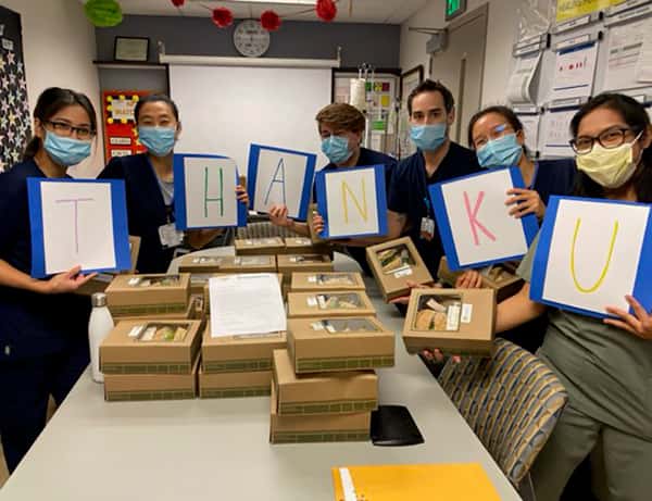Six nurses with masks standing around table full of Urth lunch boxes each holding a large card with a letter on it together spelling T H A N K U.