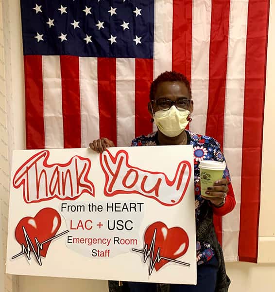 One USC nurse wearing yellow medical mask holding a sign with two big red hearts that reads: Thank You! From the HEART LAC + USC Emergency Room Staff. She is also holding a green Urth Caffé coffee cup and standing in front of a vertical USA flag.