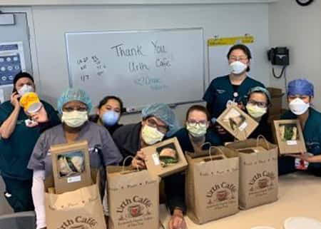 Group of nurses with masks in room with 4 Urth bags and 4 lunch boxes on table with dry erase white board behind them that say, Thank You Urth Caffé!