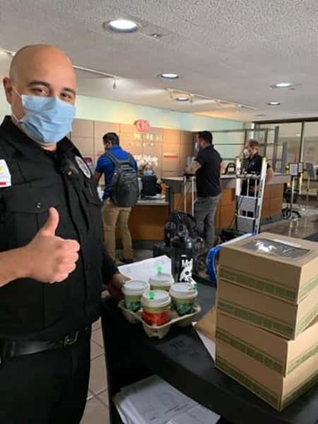 Security guard with mask standing next to cart of Urth lunch boxes and coffee cups giving thumbs up.