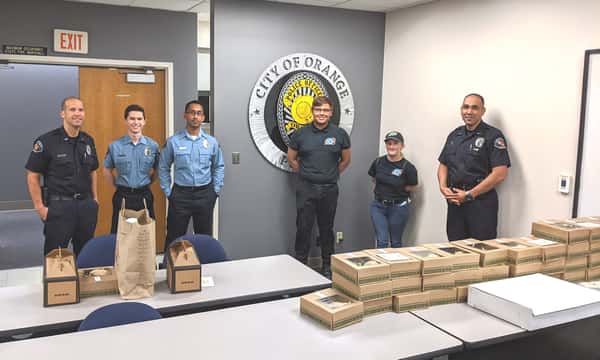 Four City of Orange policemen with two Urth employees standing in front of City seal on wall and behind a table full of Urth lunch boxes and coffee
