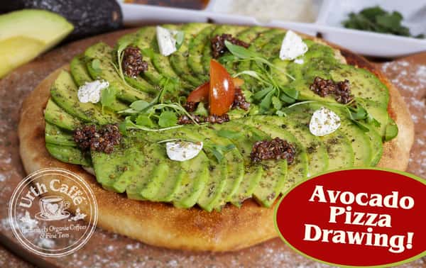 The Urth Avocado pizza on a wood pizza server with hummus, house-made tomato sauce covered with fresh sliced avocado garnished with almond cheese, micro cilantro, and quinoa. Round rustic Urth Caffé logo in the bottom left corner and an dark red oval in the bottom right saying Avocado Pizza Drawing! 
