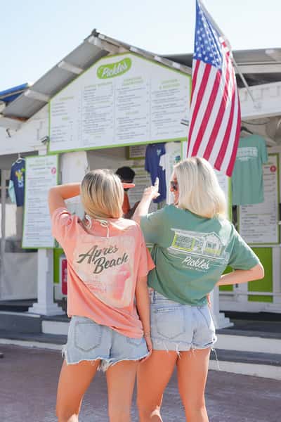 Two women in branded apparel reviewing a menu board outside a coastal restaurant, showcasing a casual beachside dining experience.