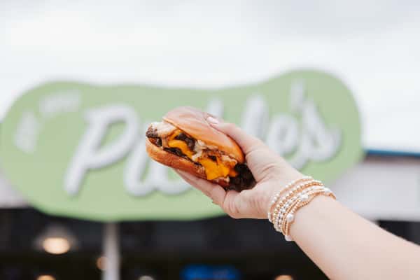 Hand holding a juicy cheeseburger in front of restaurant signage, highlighting signature beachside dining.