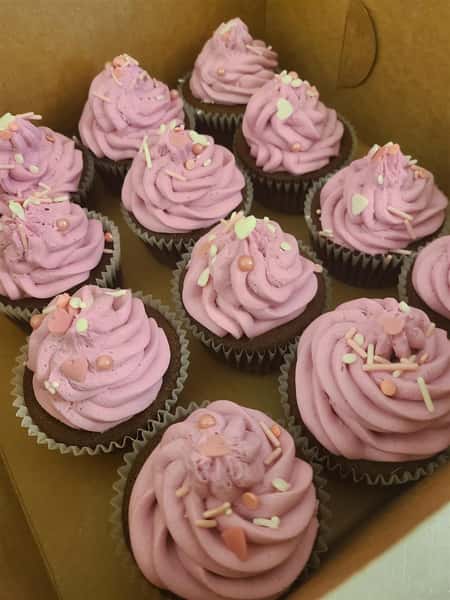 A dozen cupcakes in a box with purple frosting and sprinkles