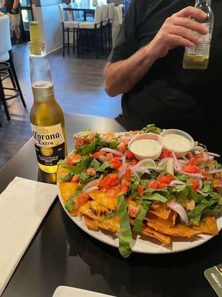A plate of nachos topped with cheese, lettuce, tomatoes, and onions, with two small containers of sour cream, and a bottle of Corona beer in the background.