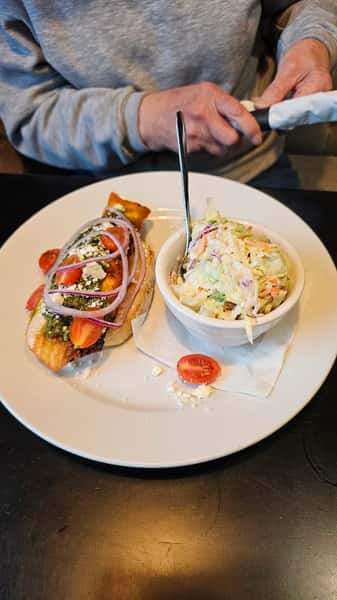 A plate featuring a grilled fish sandwich with onions, tomatoes, and pesto, accompanied by a side of coleslaw. A person is about to eat with a napkin in hand.