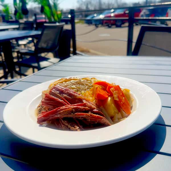 A plate of sliced roast beef served with a side of sautéed vegetables including onions, carrots, and potatoes, with outdoor seating in the background.