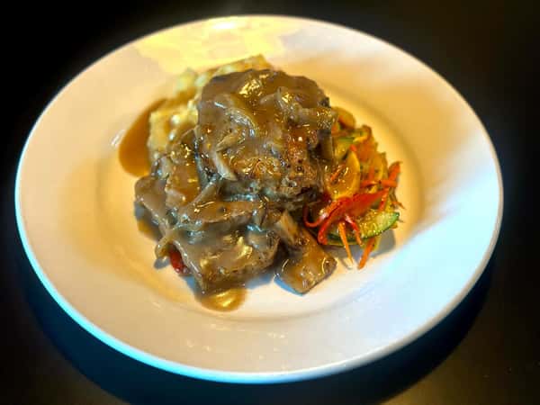 A plate with tender beef covered in gravy, served with mashed potatoes and sautéed vegetables including bell peppers, zucchini, and carrots.