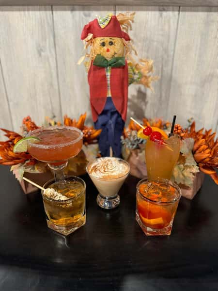 A collection of five drinks displayed on a table, including cocktails and an autumn-themed decor featuring a scarecrow surrounded by orange flowers.