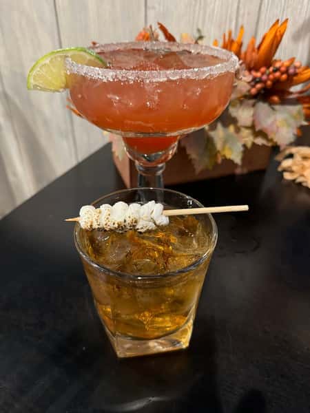 A margarita with a lime wedge and a cocktail glass with ice, topped with a skewer of roasted marshmallows.