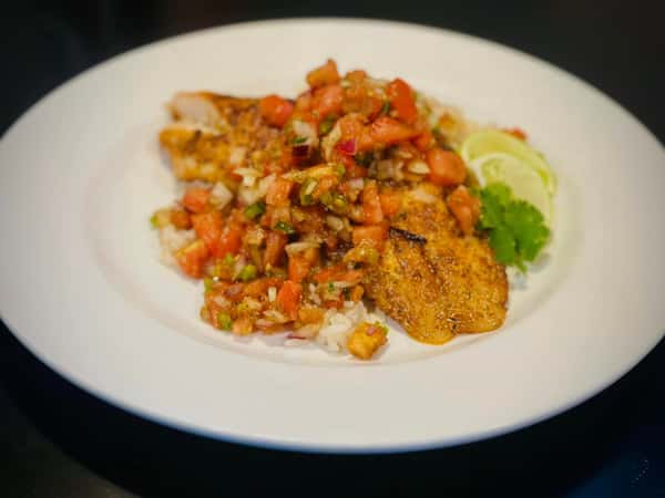 A plate of grilled fish topped with fresh tomato salsa, served over rice with lime wedges and cilantro garnish.