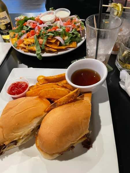 A plate with two sandwiches filled with beef, served with crispy potato wedges, dipping sauce, and a side of nachos topped with lettuce, tomato, and onions.