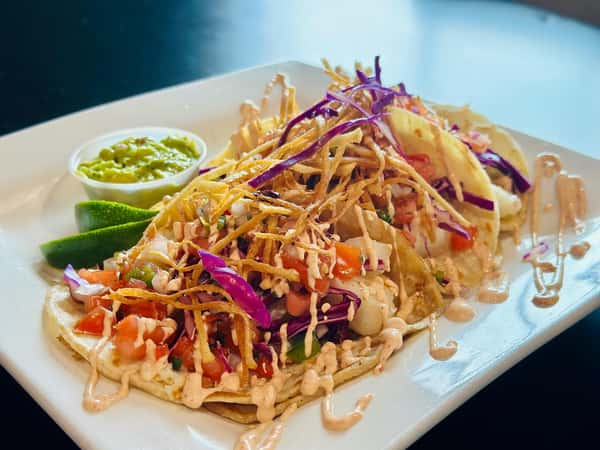 A plate with three tacos topped with shredded cabbage, crispy fried onions, tomatoes, a drizzle of creamy sauce, and served with a side of guacamole and lime wedges.