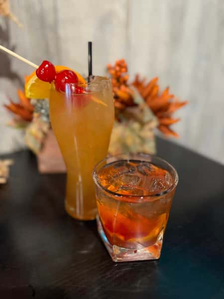 A tall cocktail with a cherry and orange garnish alongside a shorter glass with a dark drink and a cherry at the bottom.