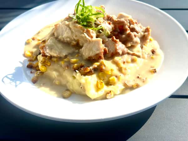 A plate of mashed potatoes topped with creamy gravy, grilled chicken, and roasted corn, garnished with green onions. The dish is served outdoors in bright sunlight.