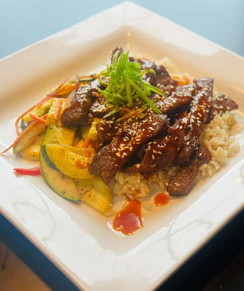 A plate of sesame beef strips served over rice with stir-fried vegetables, topped with green onions and sesame seeds.