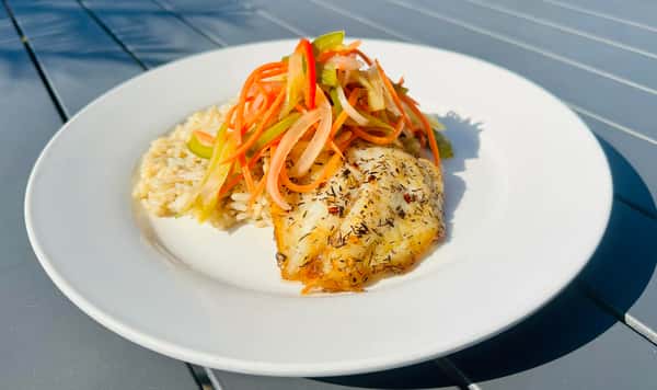 A plate with seasoned fish fillet, topped with a tangy mixture of pickled vegetables, served with rice. The dish is set on a light-colored table.