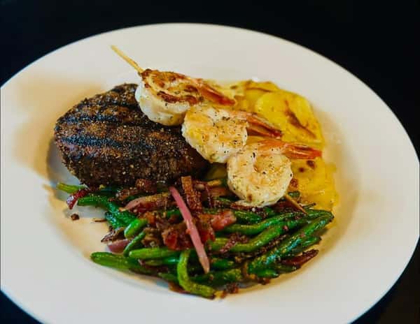 A plate featuring a grilled steak with shrimp skewers, green beans with bacon, and sliced potatoes.