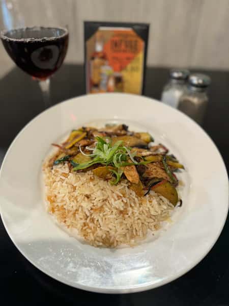 A dish of rice topped with grilled vegetables, including zucchini and bell peppers, garnished with green onions. A glass of red wine is in the background.