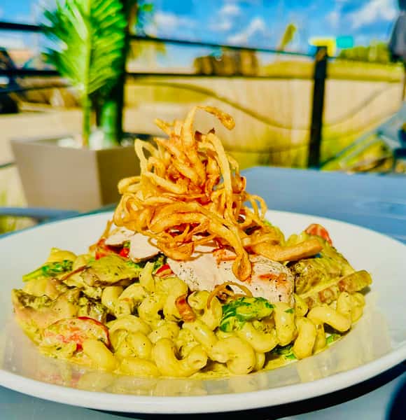 A plate of pasta with creamy pesto sauce, grilled chicken on top, and crispy fried onions, set against an outdoor background.