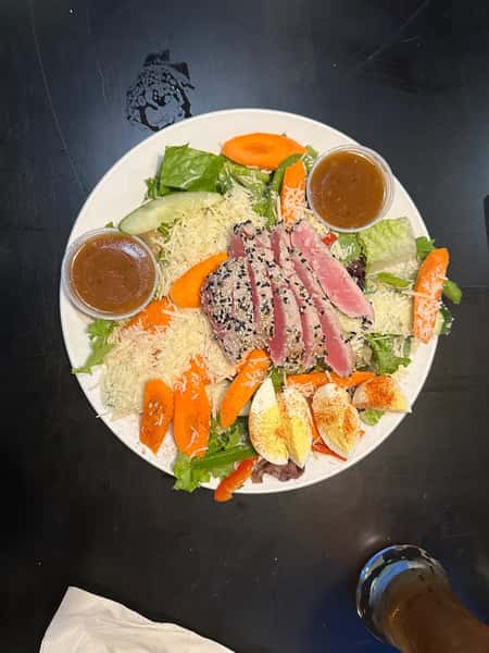 A fresh salad topped with seared tuna, carrot slices, lettuce, cucumbers, and a hard-boiled egg, served with two cups of dressing on the side.