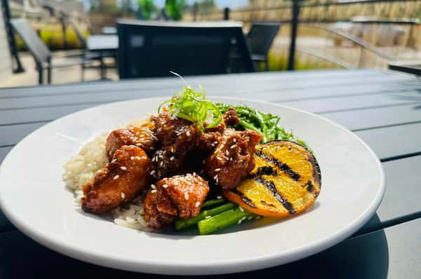 A dish featuring sesame chicken served with rice, grilled vegetables, and a charred orange slice, set on a table with an outdoor view.