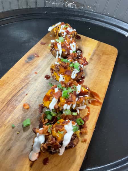 Wooden board of crispy appetizer bites topped with melted cheese, bacon, green onions, and drizzle sauce, presented as a shareable bar appetizer.