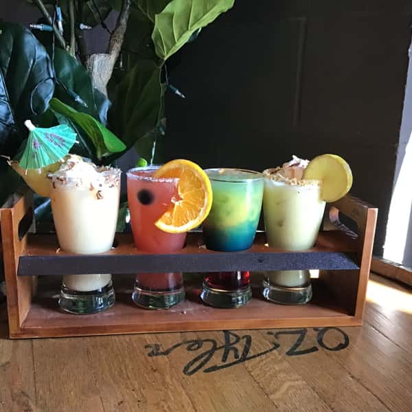 Colorful cocktail flight featuring four mixed drinks with citrus garnishes and whipped cream, served on a wooden tray in a lively bar setting.