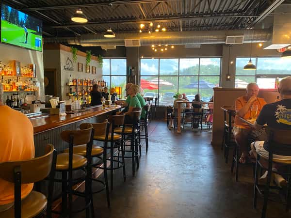 Modern restaurant interior with full bar, high-top seating, exposed ceilings, warm lighting, and guests enjoying drinks and food