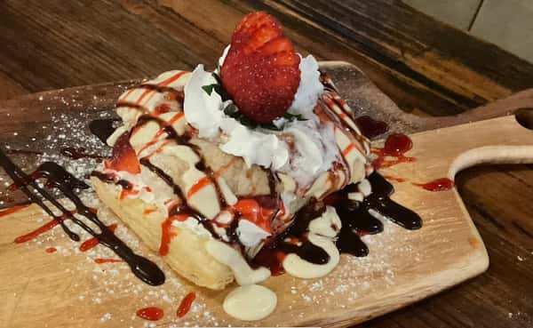 Strawberry-topped dessert pastry drizzled with chocolate and strawberry sauce, finished with whipped cream and powdered sugar on a wooden serving board.