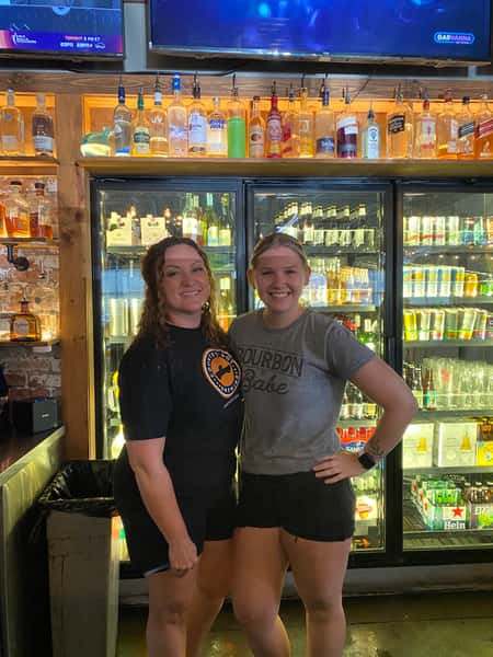 Two smiling bartenders standing in front of a backlit bar cooler stocked with beer and spirits, highlighting friendly restaurant staff.
