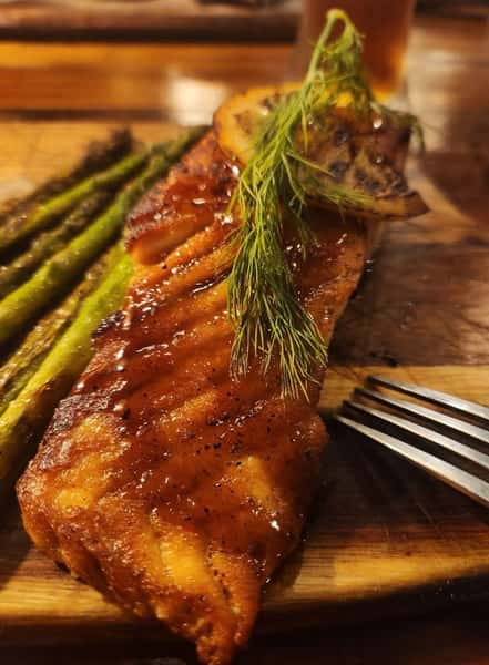 Glazed salmon fillet topped with fresh herbs and grilled citrus, served with asparagus on a wooden board in an upscale restaurant presentation.