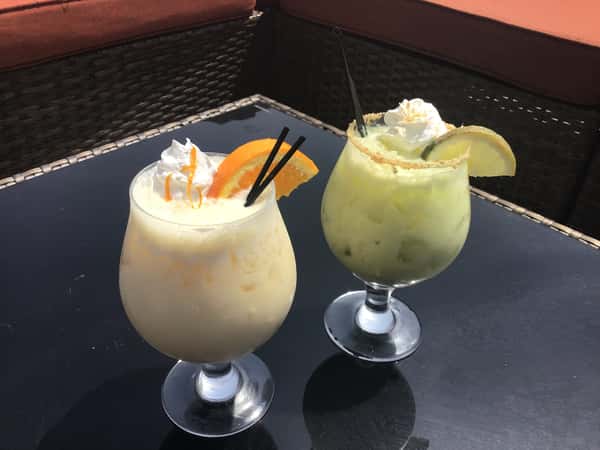 Two creamy frozen cocktails garnished with whipped cream, citrus slices, and sugar-rimmed glasses, served on a patio table at a casual restaurant.