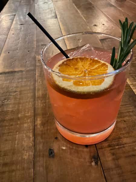 Pink citrus cocktail garnished with a dried orange slice and fresh rosemary, served over ice on a rustic wooden table.