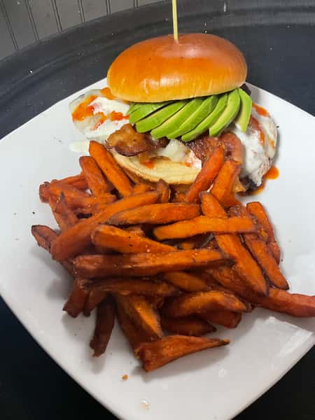 Gourmet burger topped with avocado, bacon, melted cheese, and sauce, served with crispy sweet potato fries on a white plate.