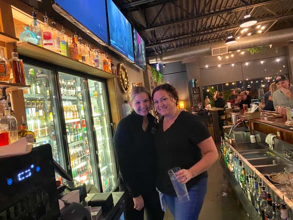 Two bartenders posing behind a full-service bar with illuminated liquor shelves and TVs, showcasing a lively bar environment