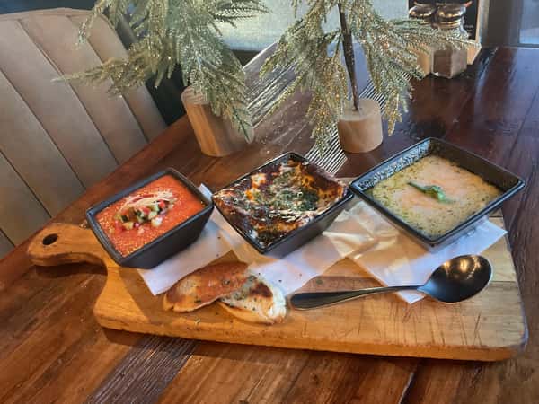 Three assorted soups with toasted bread and spoon, presented on a rustic wooden board for a comfort-food dining experience.